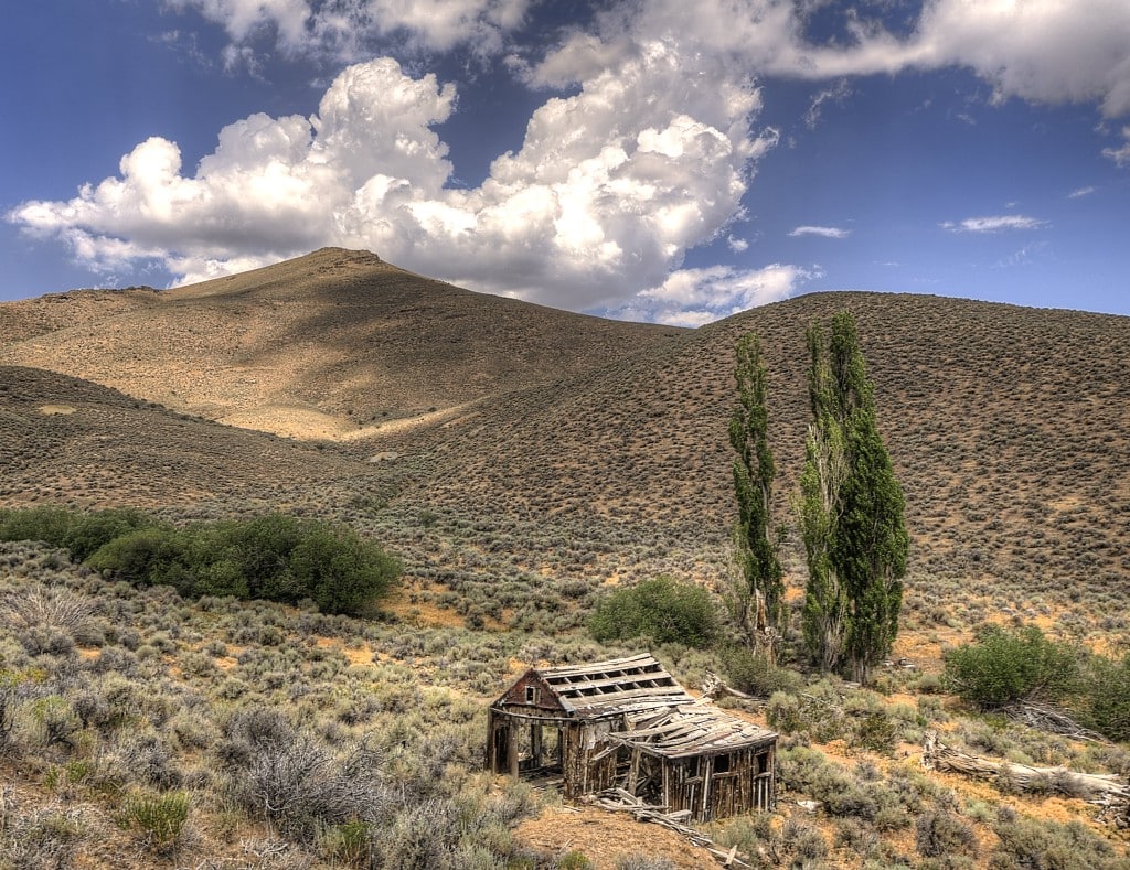 A landscape photograph showing large hills covered in desert sagebrush, with an abandoned wooden structure in the foreground, in disrepair.