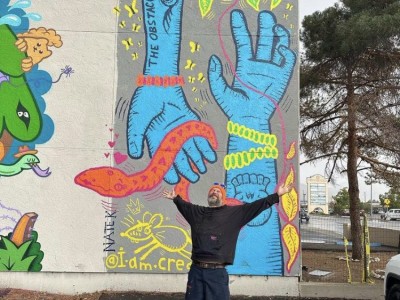 A photo of artist Nathanael Kuhn standing in front of a mural he worked on. His arms are spread wide.