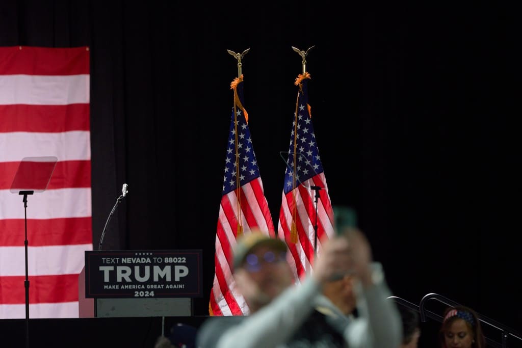 A photo showing two American flags, with a man taking a selfie in front of a stage.