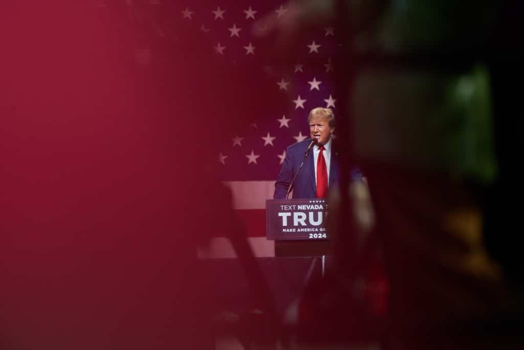 A photo of former president Donald Trump speaking behind a podium, with the foreground obscured by red and black silhouettes