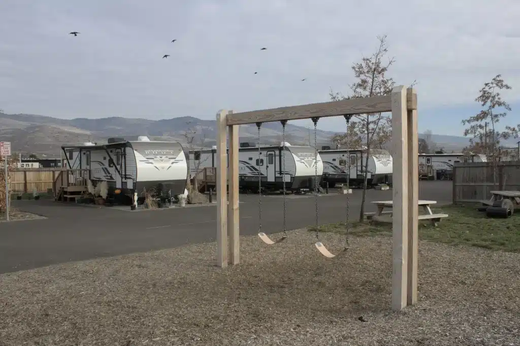 A photograph of a small swing set in front of several trailers in an outdoor community park.