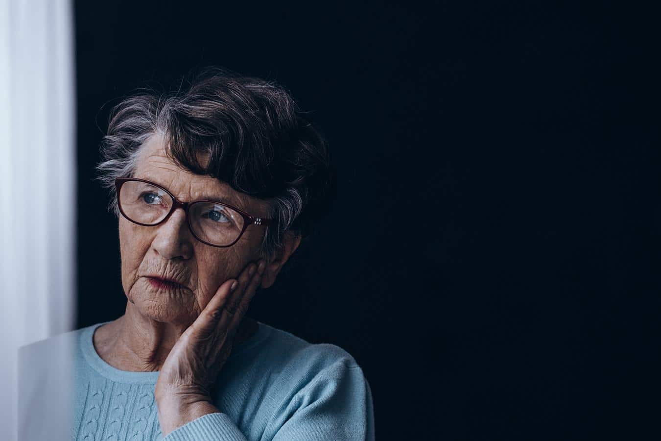 An older woman looks outside the window with a sad look on her face